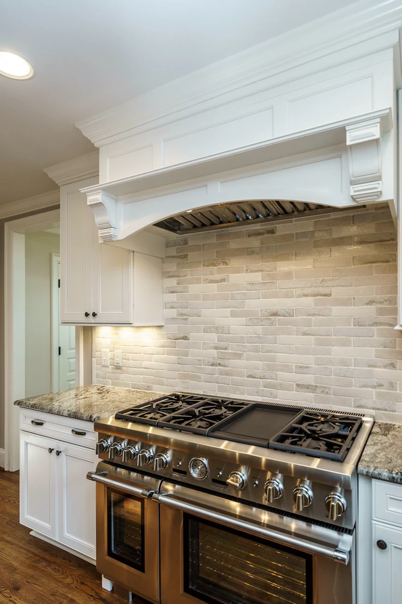 Stainless pro range with a center griddle under an arched white wood hood with corbels, light brick-style tile backsplash